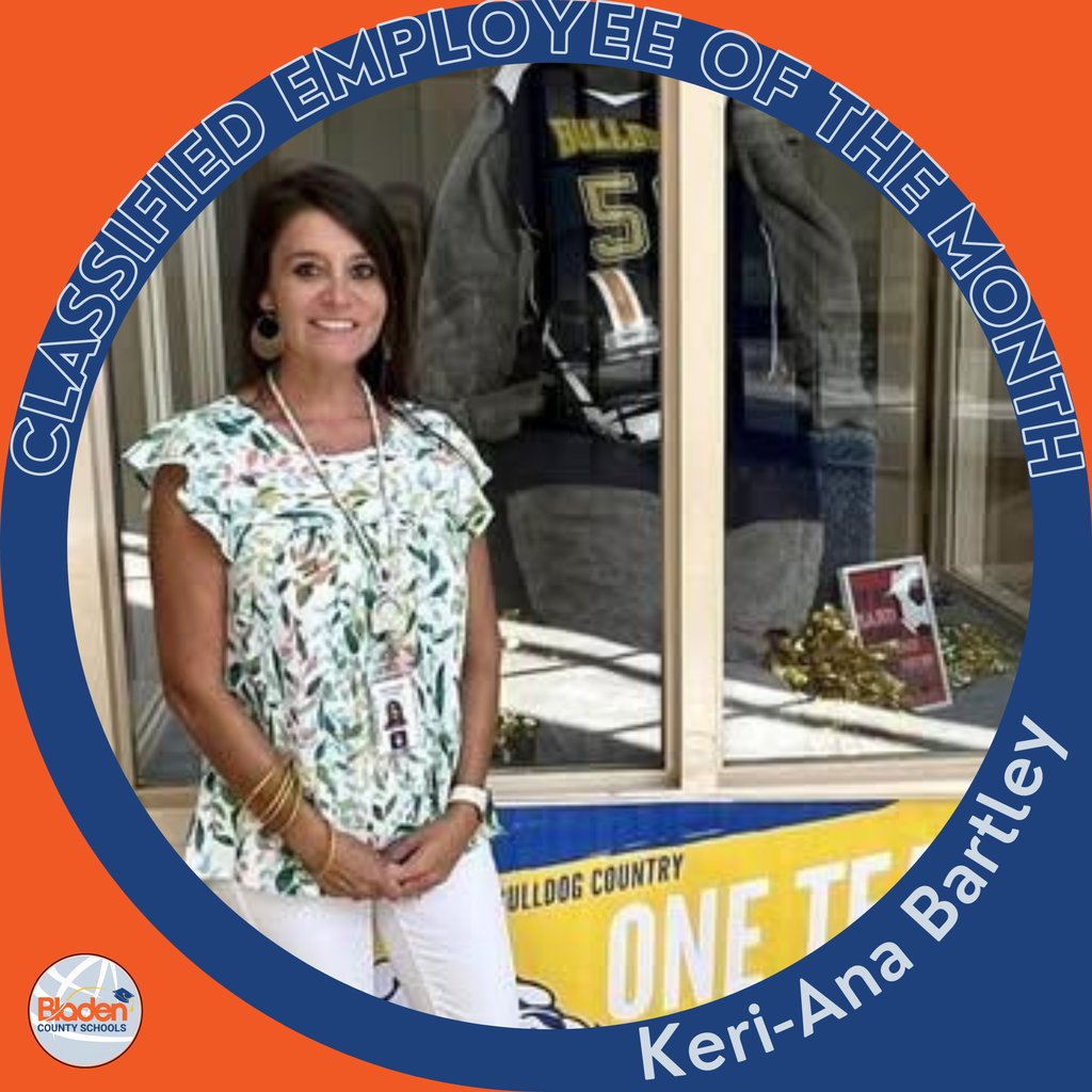 Circular ‘Classified Employee of the Month’ graphic featuring a woman smiling while standing in front of a school display case containing sports items. She is wearing a floral top with white pants and an ID badge. Text on the graphic reads ‘Keri-Ana Bartley.