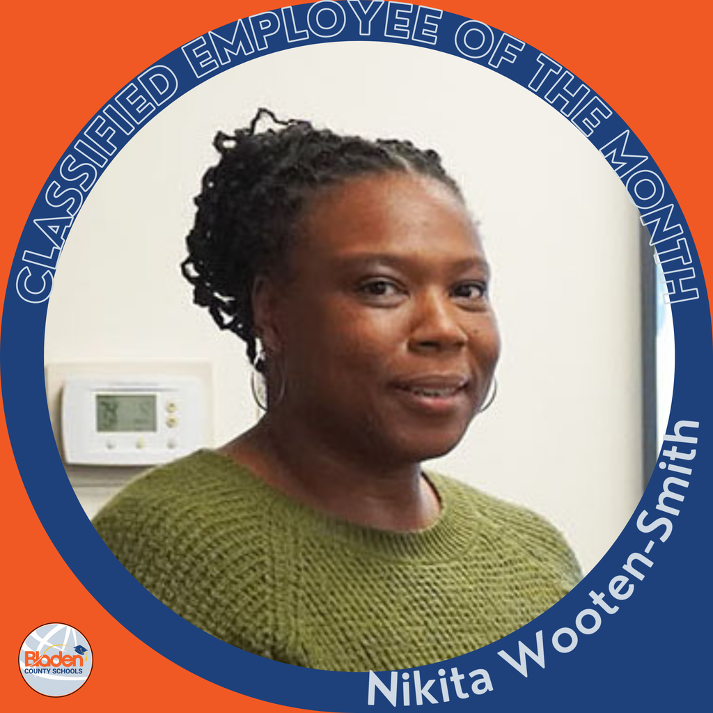 A woman wearing a green sweater smiles while standing indoors near a thermostat on the wall. Her hair is styled in braids pulled back. The circular frame reads ‘Classified Employee of the Month – Nikita Wooten-Smith,’ with the Bladen County Schools logo in the corner.