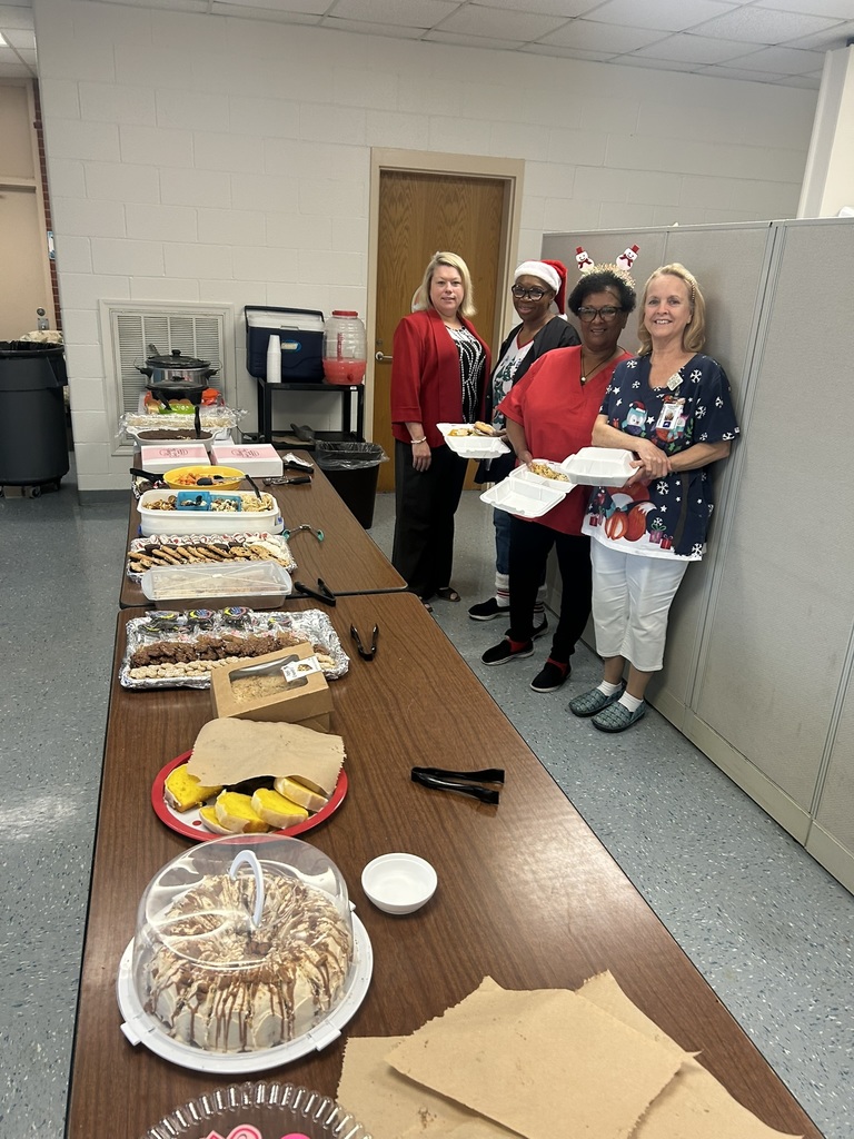 A long table is covered with a variety of holiday potluck dishes including desserts, cookies, fruit, and casseroles. At the far end of the table, four staff members stand smiling and holding plates of food. One person is wearing a Santa hat, another has Christmas-themed headbands, and all are dressed in festive holiday attire. The room appears to be a school break room with neutral-colored walls and a door in the background.