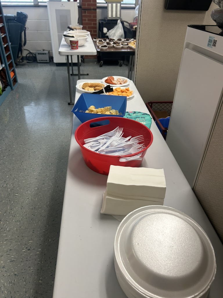 A table set up with white paper plates, napkins, plastic utensils, and small trays of crackers, cheese, and other snacks. More potluck food can be seen on additional tables further back, including dips, cupcakes, and assorted dishes. The scene is inside a school setting with gray flooring and cubicle dividers visible.