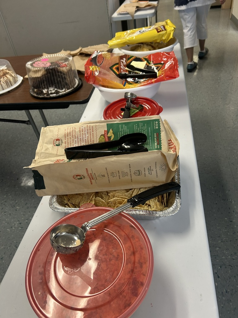 A close-up view of part of the potluck setup. Bowls and trays of chips, dips, and toppings sit on a white table. A large bag of tortilla chips, serving spoons, and a covered tray of crumbled toppings are visible. A chocolate cake in a plastic dome sits on the neighboring table. A staff member’s legs and Christmas-themed scrubs are partially visible walking away.