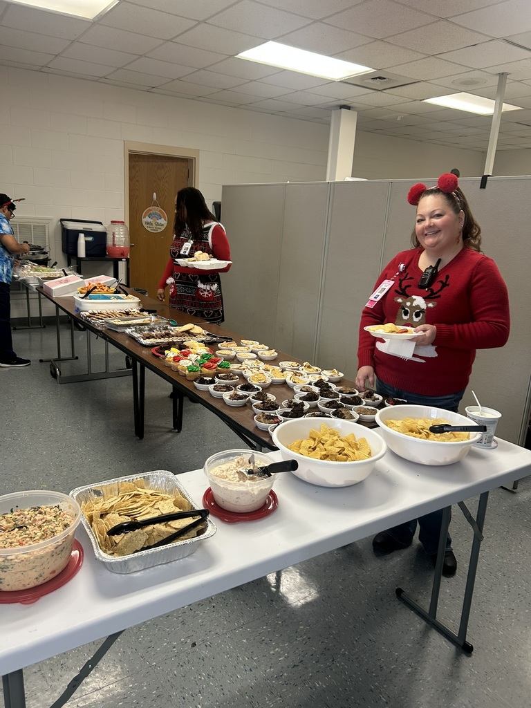 nother long table is filled with decorated holiday cupcakes, cookies, chips, dips, and other potluck dishes. A staff member wearing a red sweater with a reindeer design and fuzzy red Christmas headband stands smiling with a plate of food. Another person in a festive dress stands nearby looking at the food. More tables of food extend into the background.