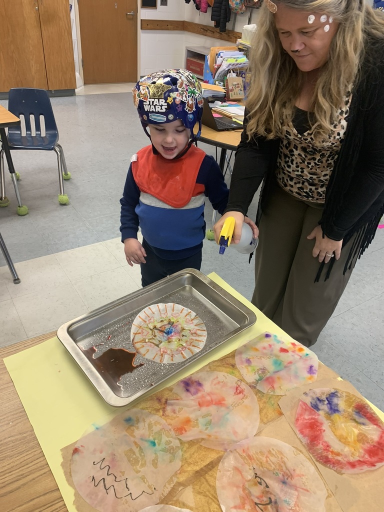 A teacher assists a student as they spray water onto a coffee-filter snowflake, watching the colors blend and flow across the paper.