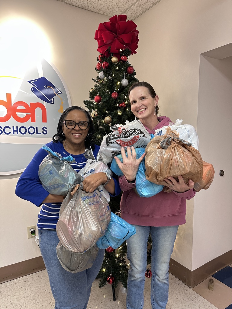 Two staff members smile while holding several bags of donated food in front of a decorated Christmas tree inside a school building. The Bladen County Schools logo is visible on the wall behind them.