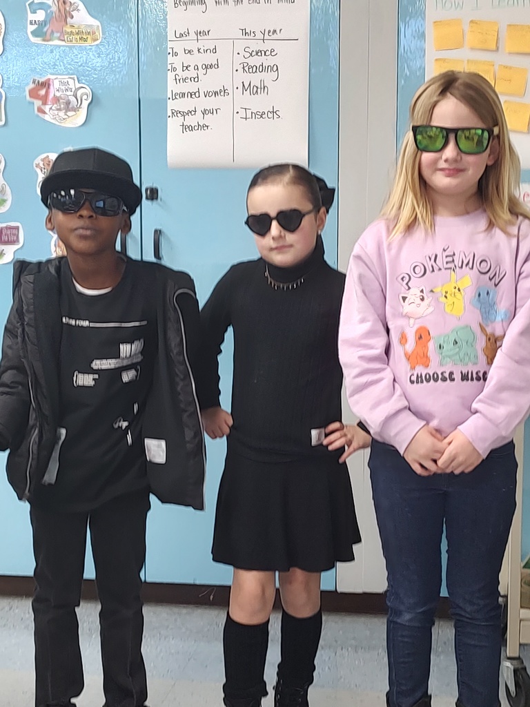 Three elementary students stand side by side indoors, dressed for “I Spy Santa” dress-up day. All three are wearing sunglasses and winter outfits. One student wears an all-black outfit with a jacket and hat, another wears a black dress with heart-shaped sunglasses, and the third wears a pink Pokémon sweatshirt with green sunglasses. They pose confidently in front of a light blue classroom wall with student work displayed.
