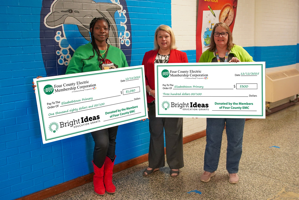 Three EPS staff members stand in a school hallway holding large Bright Ideas Education Grant checks from Four County Electric Membership Corporation. Asia Burney holds a check for $1,080 and Caren Long holds a check for $300.