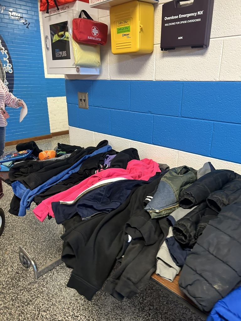 ables in the front lobby of Elizabethtown Primary School filled with lost and found items including coats, jackets, hoodies, and sweaters in various colors.