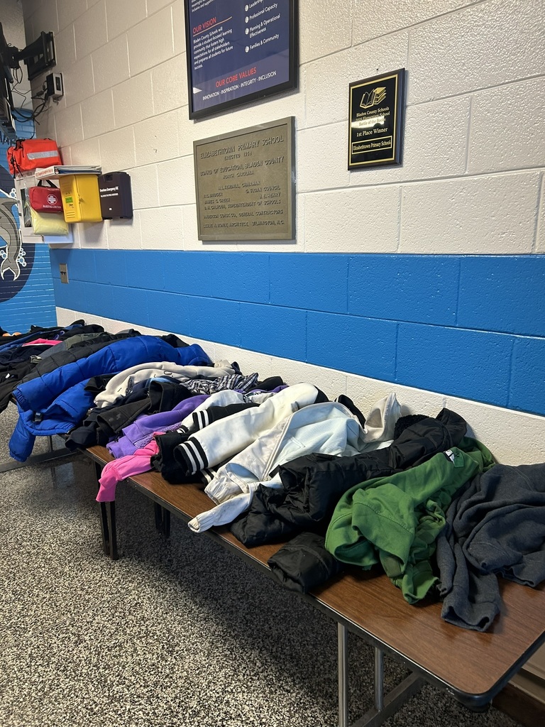 ables in the front lobby of Elizabethtown Primary School filled with lost and found items including coats, jackets, hoodies, and sweaters in various colors.