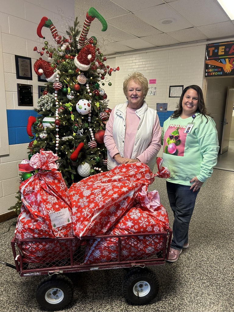 Two adults stand beside a decorated Christmas tree in a school hallway next to a wagon filled with wrapped Christmas gifts. The gifts were donated by Operation Blessing Bladen’s Children, representing Camp Clearwater Chapel in White Lake, to support students at Elizabethtown Primary School.