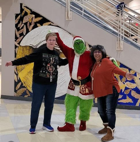 Two staff members pose with the Grinch in a school lobby area with a large mural visible in the background.