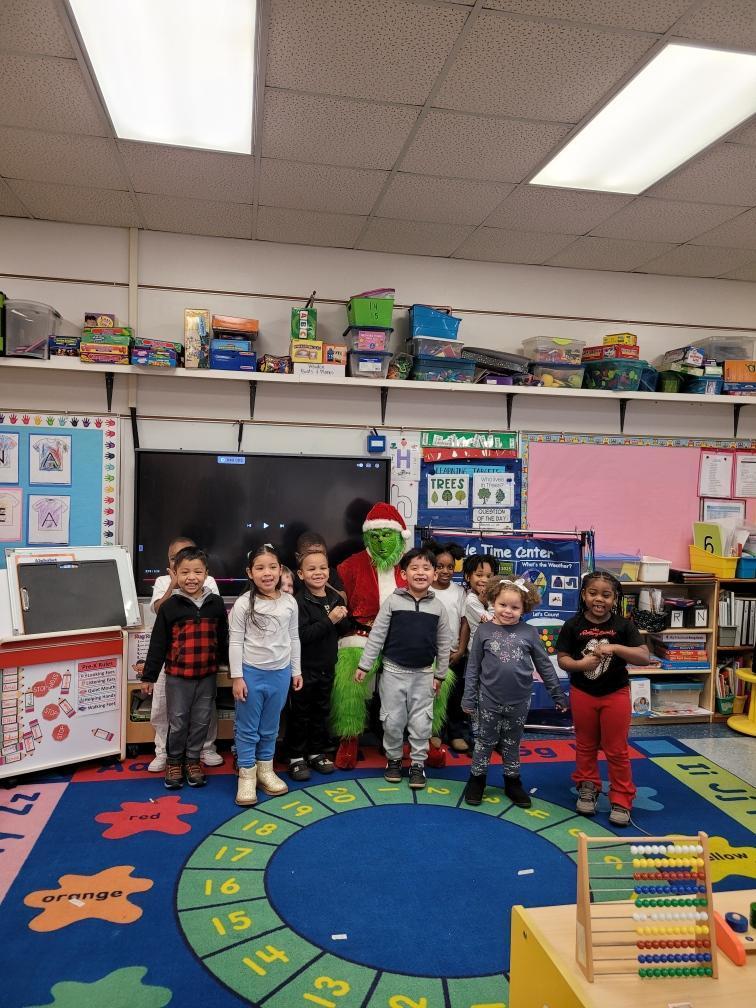A group of young students stand on a classroom rug smiling while posing with the Grinch during a classroom holiday visit.