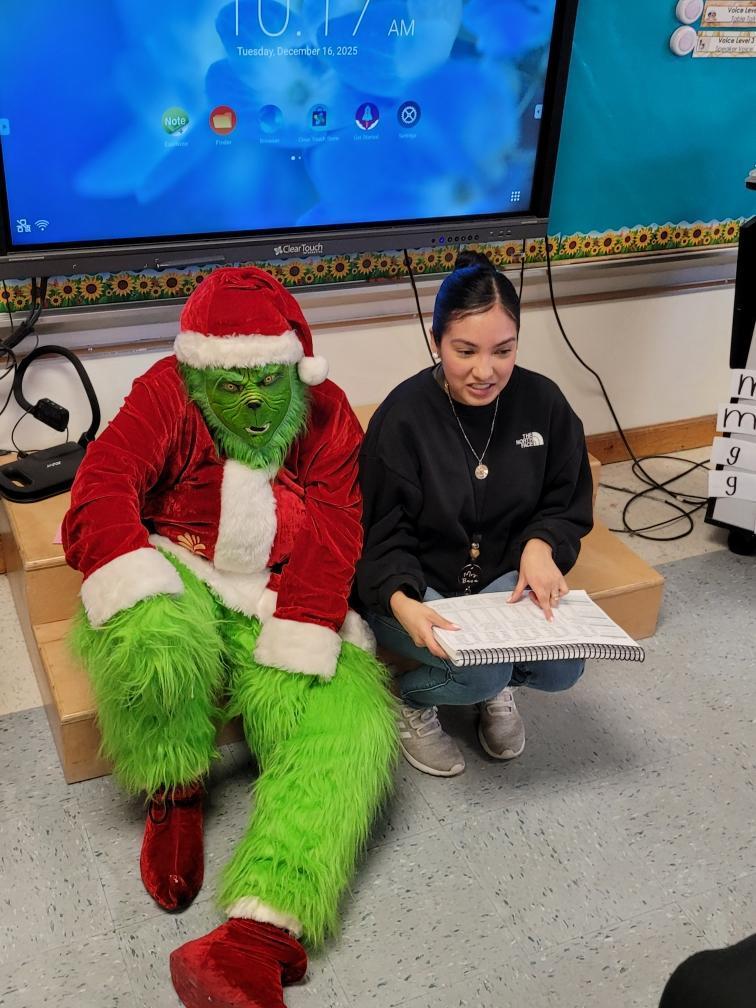 The Grinch sits on classroom steps beside a teacher who is holding a book, appearing to read aloud to students off camera.