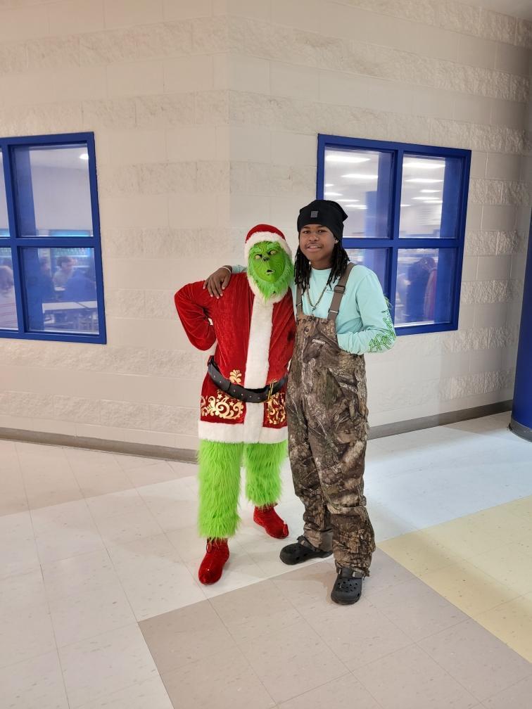 A student stands with an arm around the Grinch while posing for a photo in a school hallway.