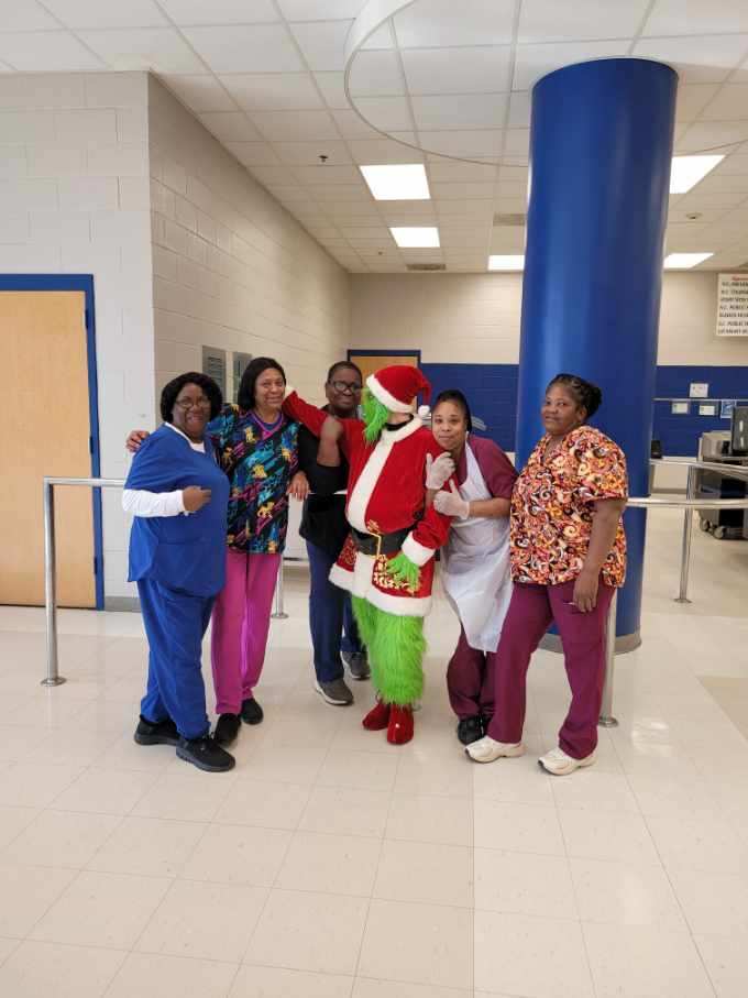 Cafeteria staff pose together with the Grinch in a school cafeteria during a holiday visit.