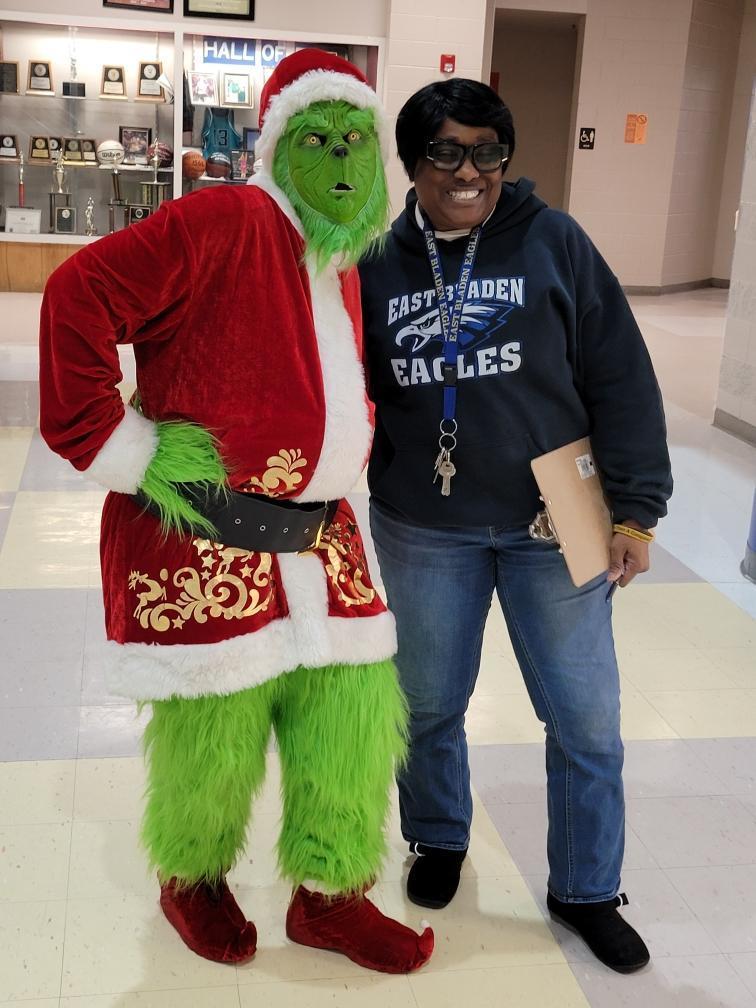 A school staff member smiles while posing in a hallway next to the Grinch during a holiday-themed school visit.