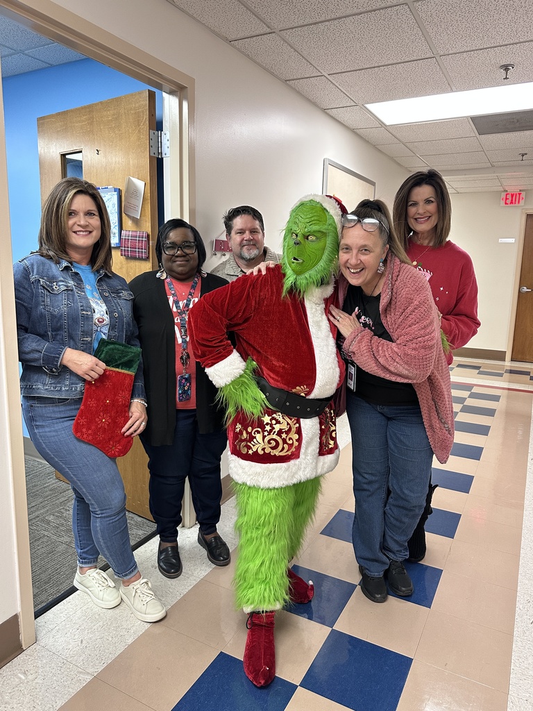 A group of school staff members pose in a hallway with a person dressed as the Grinch wearing a Santa-style outfit during a holiday visit.