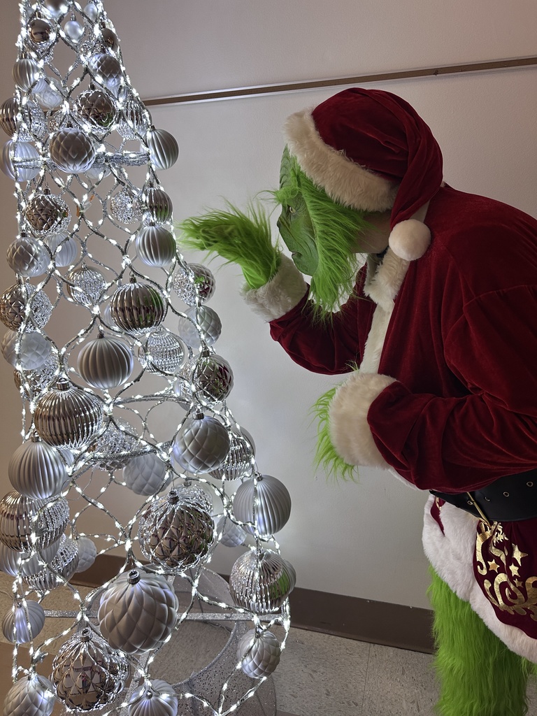 The Grinch costume character leans toward a silver holiday tree decorated with metallic ornaments and white lights in a school hallway.