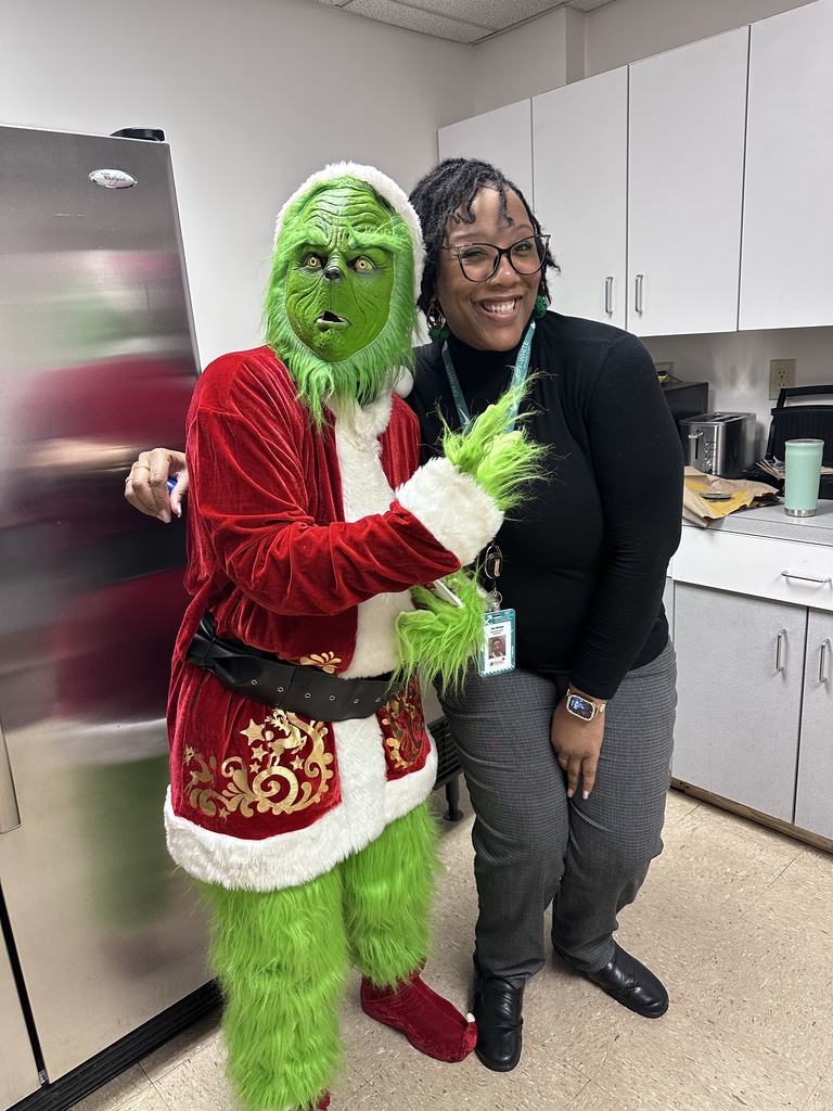A staff member smiles while posing with a person dressed as the Grinch in a school staff break room with cabinets and appliances visible.