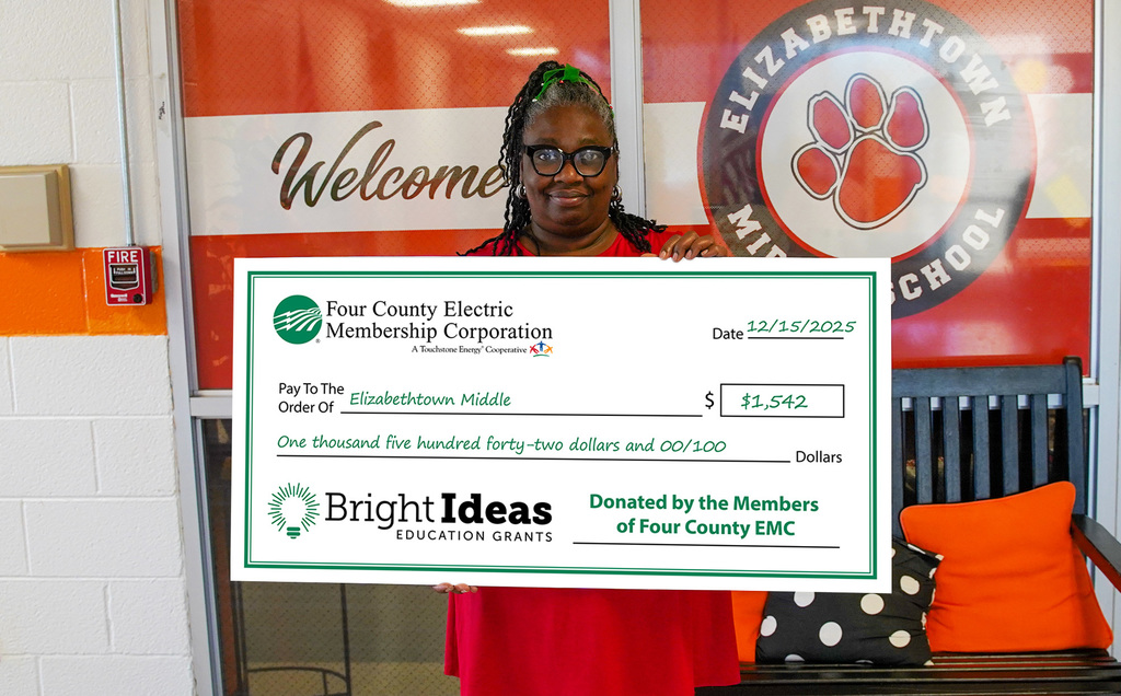 A smiling staff member stands inside Elizabethtown Middle School holding a large ceremonial check for $1,542 from Four County Electric Membership Corporation’s Bright Ideas Education Grants program, awarded to support Elizabethtown Middle School, with the school logo and orange décor visible in the background.