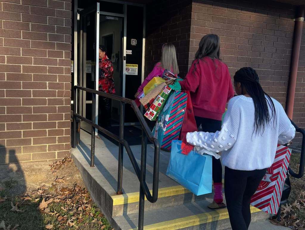Four elementary-aged students carry large holiday gift bags as they enter a school building through an exterior doorway.