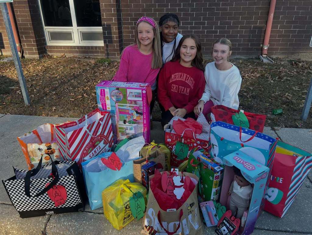 Four elementary-aged students sit outside a school building surrounded by donated holiday gifts and colorful gift bags.