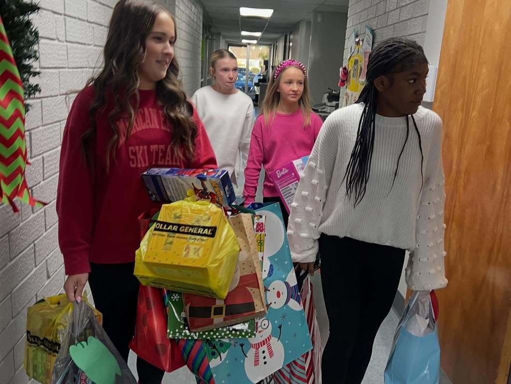Four elementary-aged students walk down a school hallway carrying donated holiday gifts and bags.