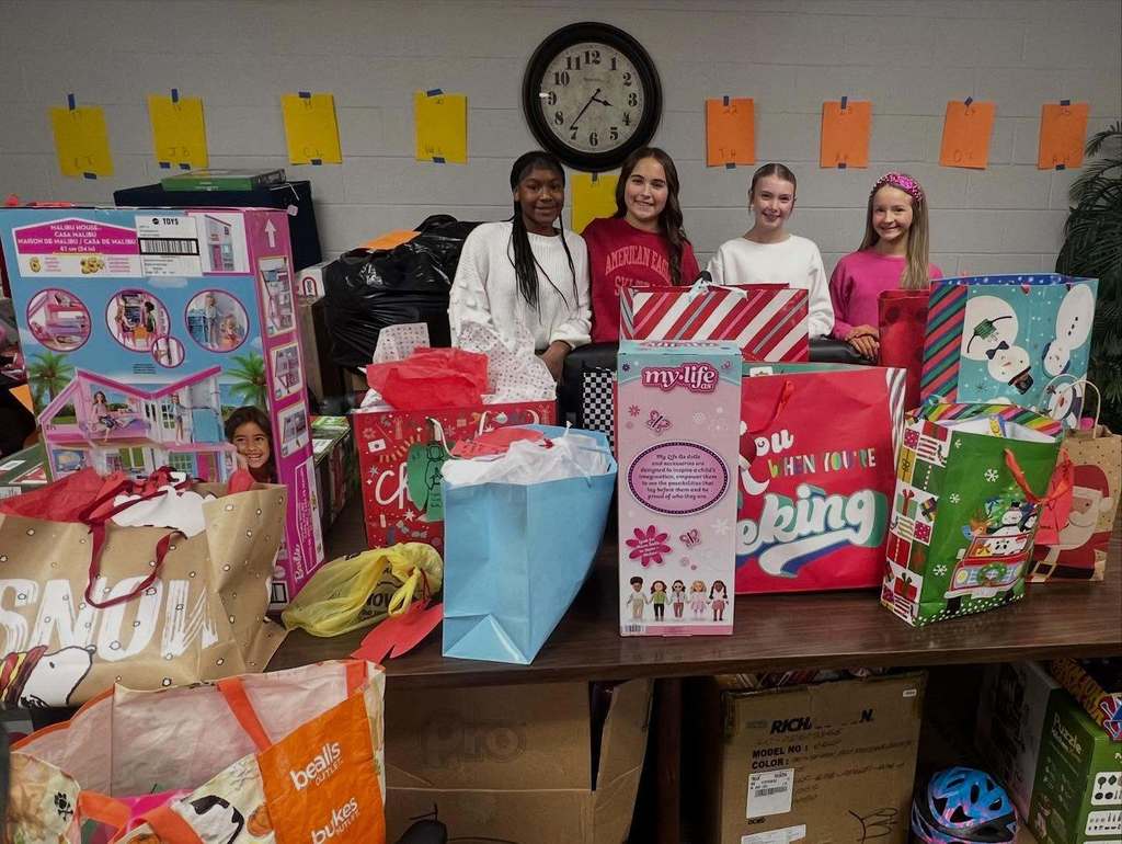 Four elementary-aged students stand behind a table filled with donated holiday gifts and gift bags inside a school building.