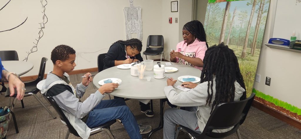 Students sit around a classroom table working collaboratively on a hands-on science activity, carefully examining materials on paper plates with tools, while educational wall art and a forest mural surround them.