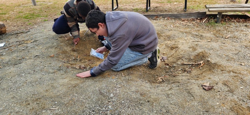 Two students kneel on the ground outdoors, closely examining soil and gravel as part of a science investigation, using tools and observation to search for small fossils or natural materials.