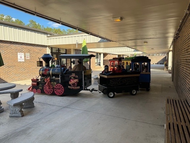 A small trackless train is parked in a covered school walkway, with students seated inside colorful train cars as part of a fun school activity or special event during the school day.