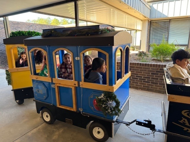 Young students smile and wave from inside a yellow train car, visible through the windows, as they enjoy a cheerful train ride during a school celebration or special activity.