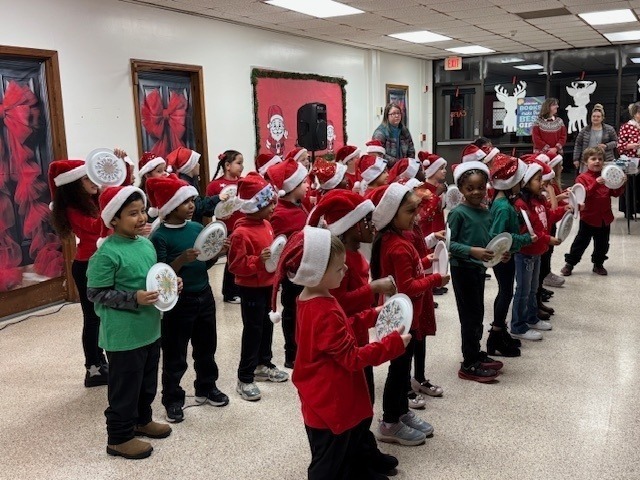 A large group of students wearing red and green outfits and Santa hats hold paper plates as props while singing together during a cheerful holiday concert at school.