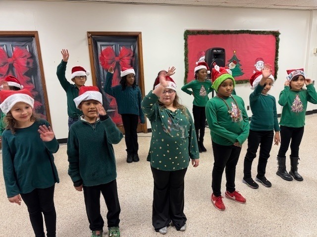 Students dressed in green shirts and Santa hats stand in rows, waving during a holiday performance in a school hallway decorated with festive artwork and seasonal displays.