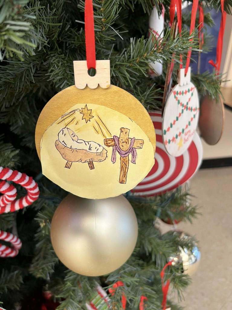 Close-up of a student-painted ornament depicting a nativity scene with baby Jesus in a manger and a wooden cross, hanging on a Christmas tree.