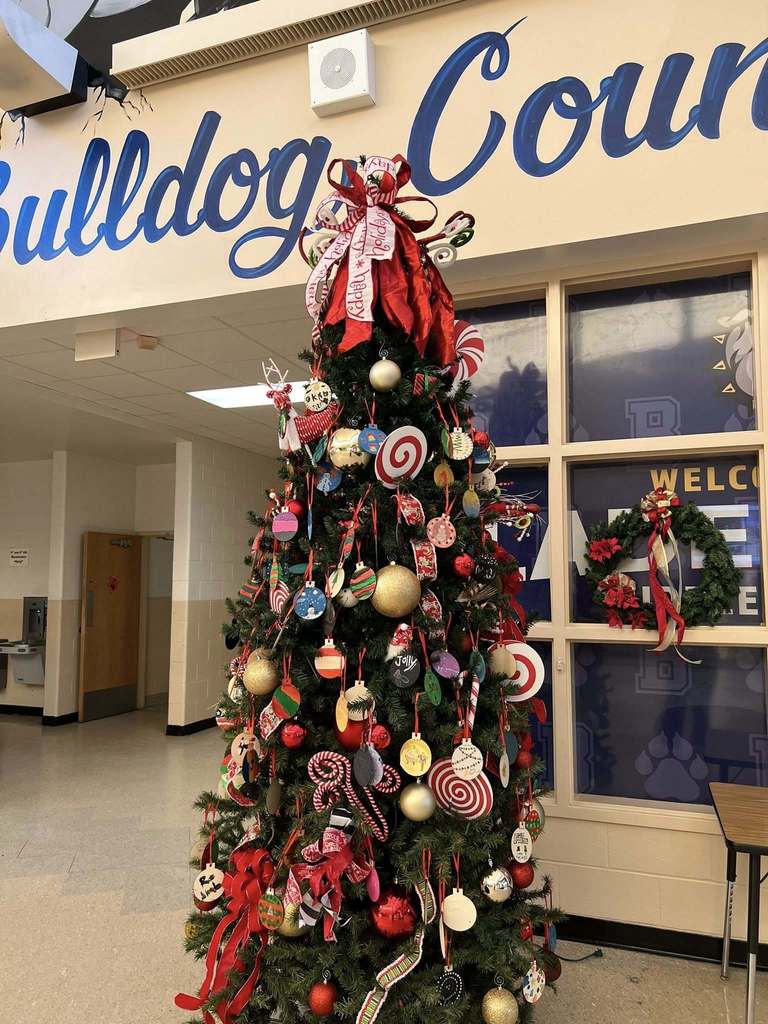 Wide view of a tall Christmas tree in a school hallway, decorated with student-made ornaments and ribbons beneath a “Bulldog Country” sign.