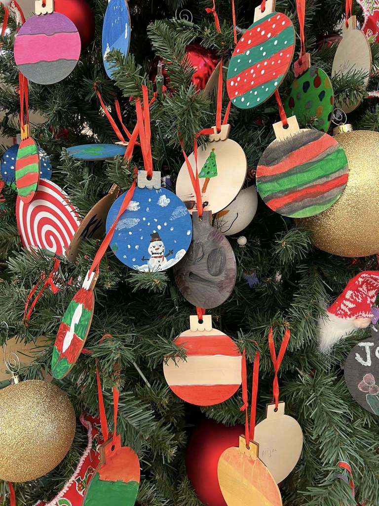Close-up of a Christmas tree decorated with student-painted wooden ornaments in festive colors, including snowmen, stripes, and patterns.