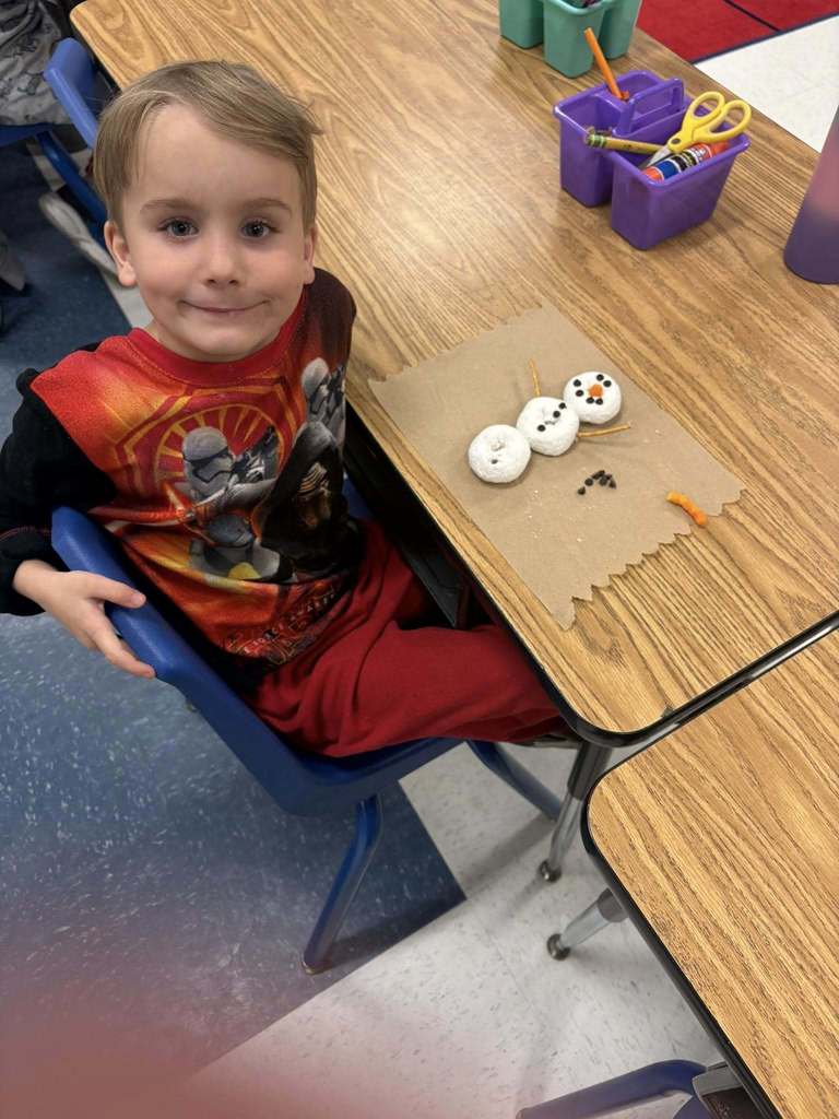 Student in red clothing sits at a desk, smiling next to a marshmallow snowman craft in progress.