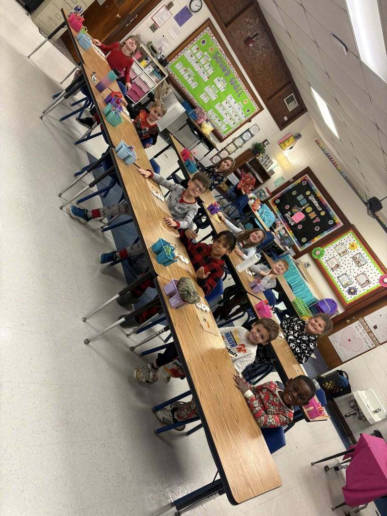 Elementary students sit at desks in a classroom, smiling while working on a holiday craft activity together.