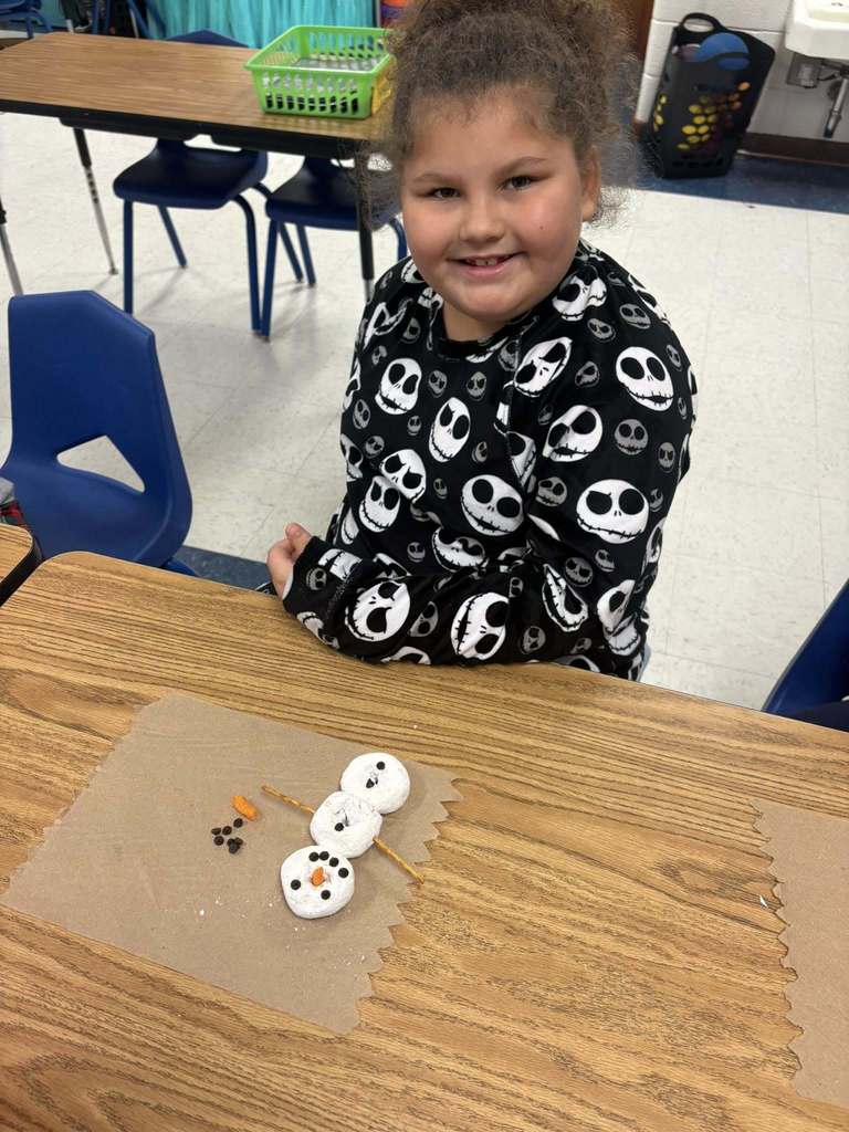 Smiling student sits at a desk wearing a black skull-pattern sweatshirt, displaying a marshmallow snowman craft.