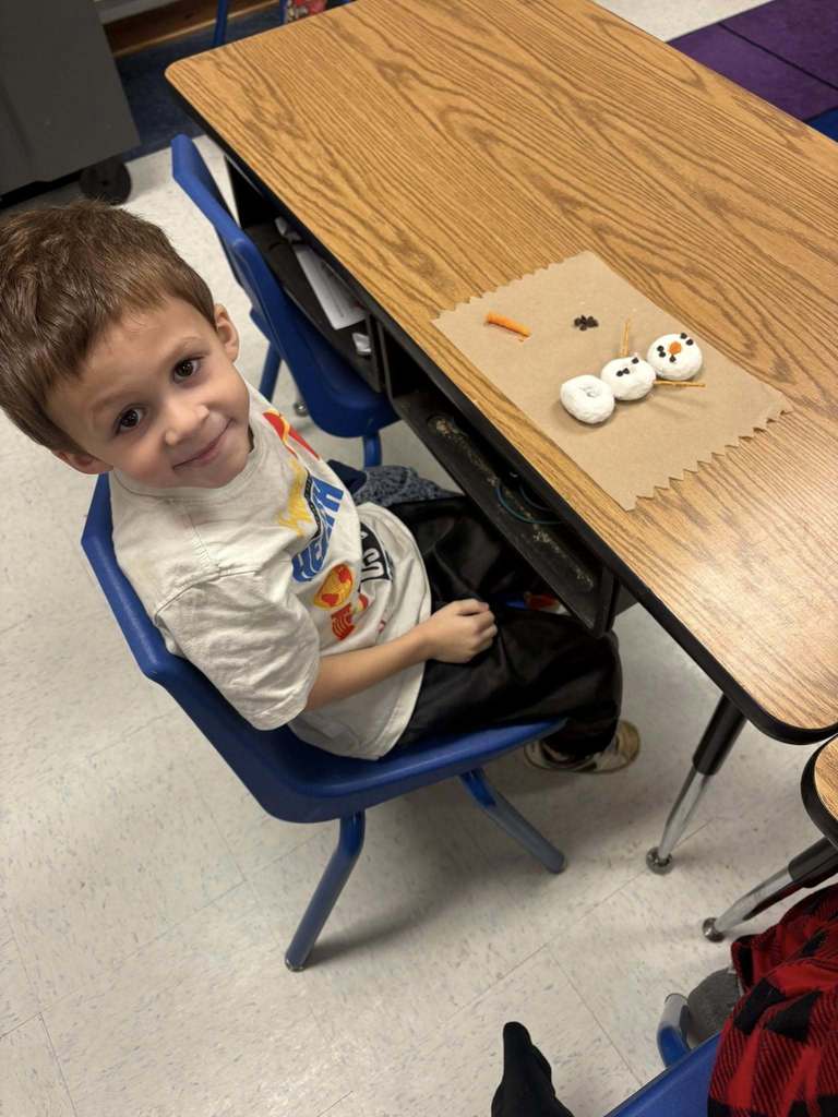 Student seated at a classroom desk smiles beside a marshmallow snowman made with pretzels and candy.