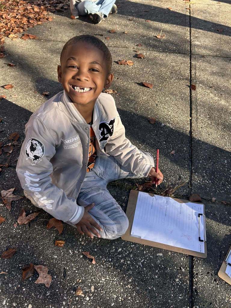 A student smiles while sitting on the sidewalk, holding a pencil and clipboard during an outdoor classroom assignment.