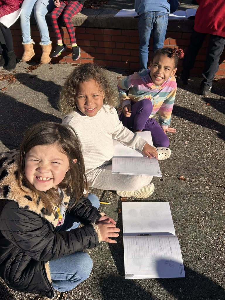 A small group of students sit on the pavement with open notebooks, smiling at the camera during an outdoor learning activity.