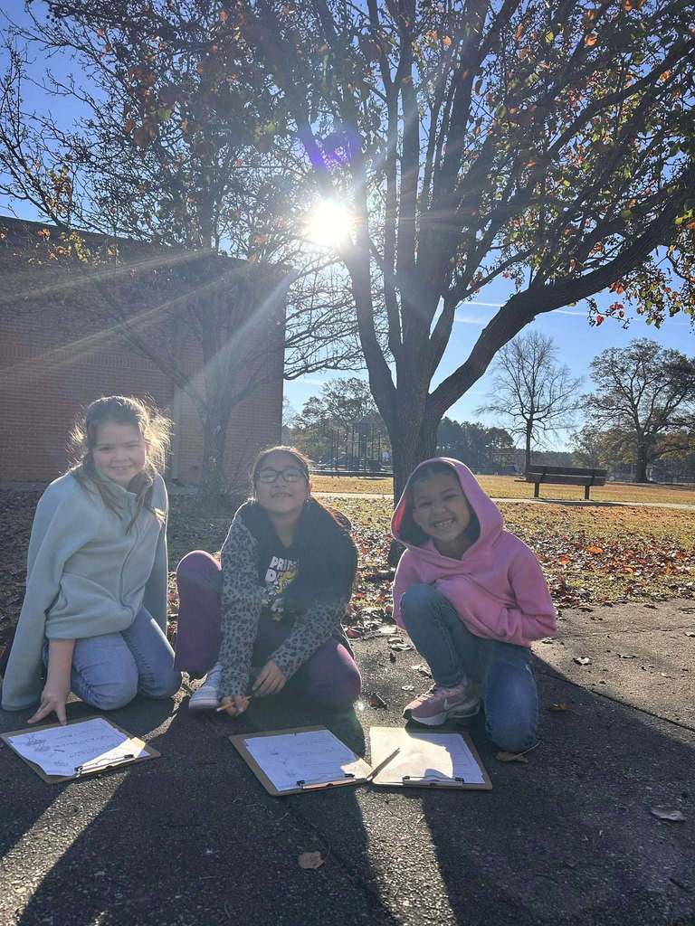 Three students kneel on a sidewalk outdoors, smiling while completing worksheets beneath a tree on a sunny fall day.