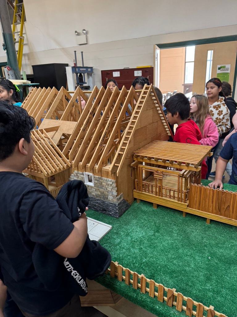 Elementary students observe a detailed wooden model of a house structure, learning about framing and construction techniques in a workshop setting.