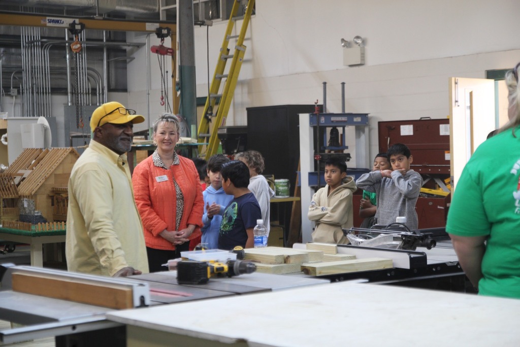 An instructor addresses elementary students gathered around a worktable with tools and materials inside a construction workshop.