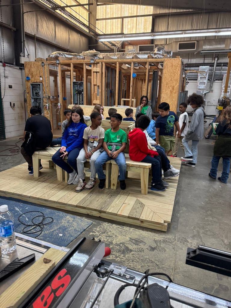 Elementary students observe a detailed wooden model of a house structure, learning about framing and construction techniques in a workshop setting.