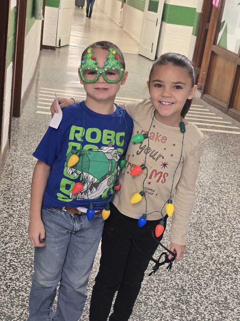 Two elementary students smile in a school hallway wearing holiday-themed accessories, including light-up necklaces and festive glasses.