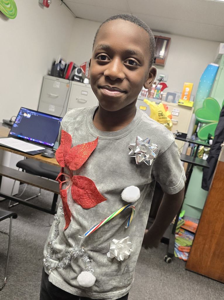 An elementary student smiles while wearing a creatively decorated holiday shirt adorned with bows, pom-poms, and festive details.