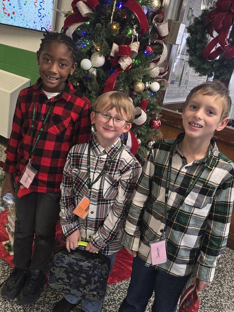 Three elementary students smile while standing in front of a decorated Christmas tree inside a school hallway.