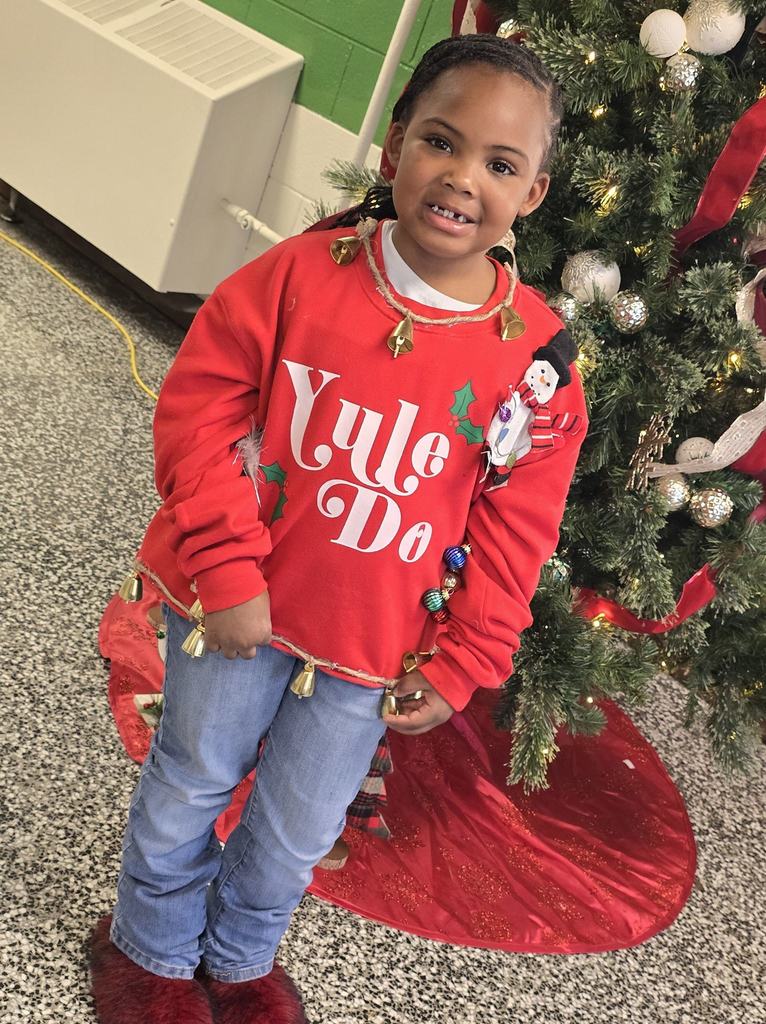 A young student wearing a festive red holiday sweatshirt poses proudly in front of a decorated Christmas tree at school.